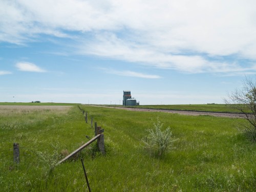 A green field stretches towards a distant grain elevator under a blue sky with scattered clouds.