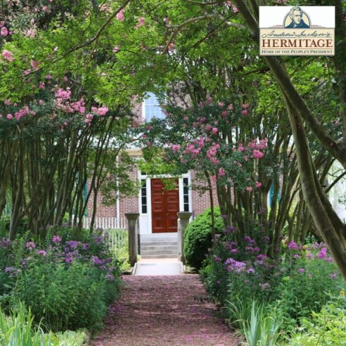 A pathway lined with blooming flowers leads to a historic brick house surrounded by lush greenery.