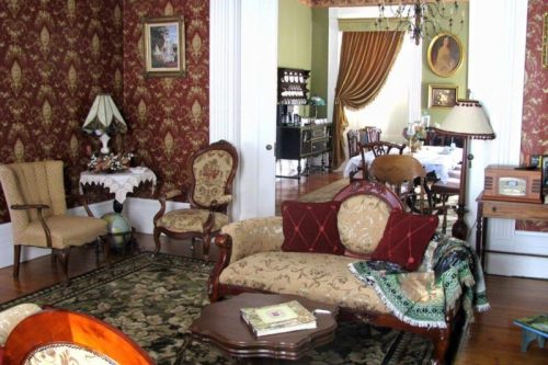 A vintage living room with ornate wallpaper, elegant furniture, and a cozy atmosphere, leading to a dining area.