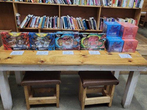 A wooden table displays various Pokémon card sets, with bookshelves filled with books in the background.