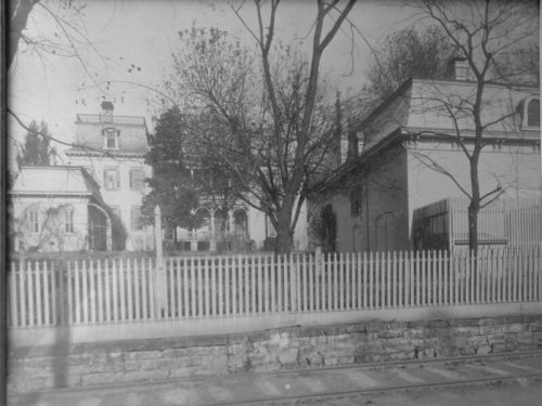 Black and white image of a large house with a picket fence, trees, and another building nearby.