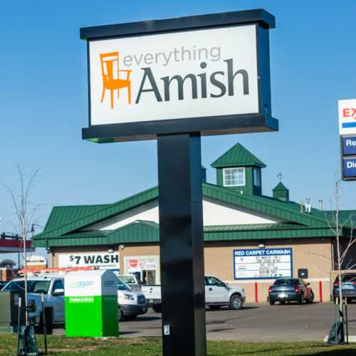 Sign for "Everything Amish" store with a chair logo, located near a car wash and other businesses. Clear blue sky.