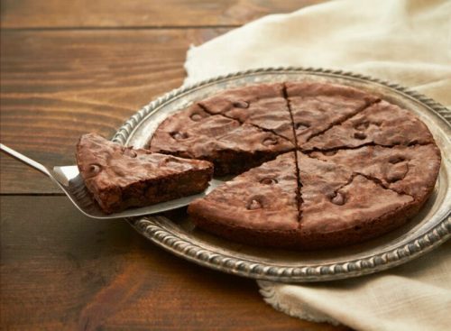 A chocolate brownie pie on a silver platter, with a slice being lifted by a spatula.