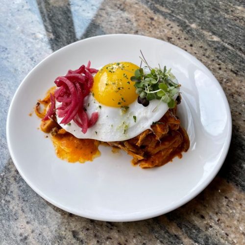 A plate of savory dish topped with a sunny-side-up egg, pickled onions, and microgreens on a marble surface.