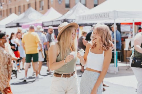 Two young women enjoy ice cream at a lively outdoor market, surrounded by colorful tents and people.