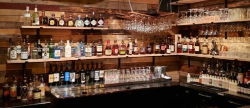 A well-stocked bar with various bottles of liquor on wooden shelves and a row of glasses hanging above.