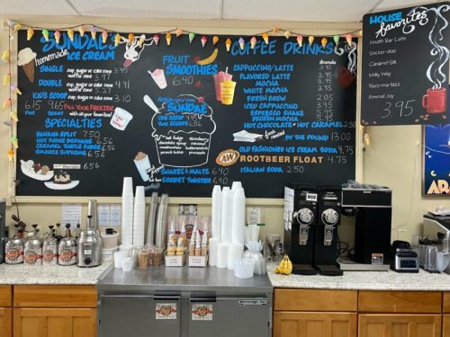 Menu board featuring ice cream, smoothies, coffee drinks, and prices, with a counter and drink dispensers below.