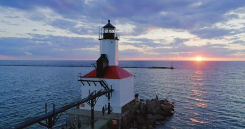 A lighthouse stands on a pier at sunset, with calm waters and a colorful sky in the background.