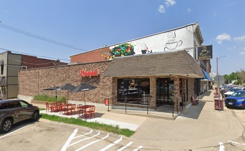 A brick café with outdoor seating, umbrellas, and a sign that says "Open," set in a sunny urban area.
