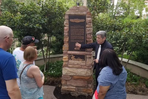 A group of people listens to a speaker by a stone monument with a plaque in a garden setting.