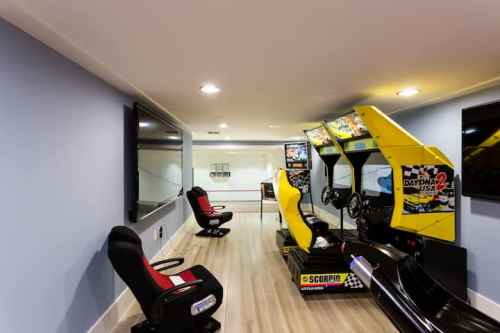 A modern game room featuring arcade racing machines, comfortable seating, and large wall-mounted TVs.