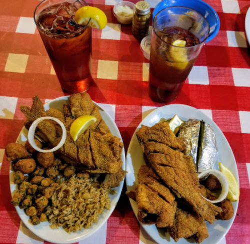 Two plates of fried fish with sides of rice, okra, and a baked potato, accompanied by iced tea in blue cups.