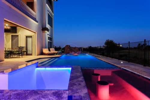A luxurious pool area at dusk, featuring a lit spa, lounge chairs, and a scenic view.
