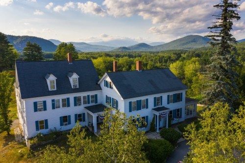 Aerial view of a white house with blue shutters, surrounded by trees and mountains under a partly cloudy sky.