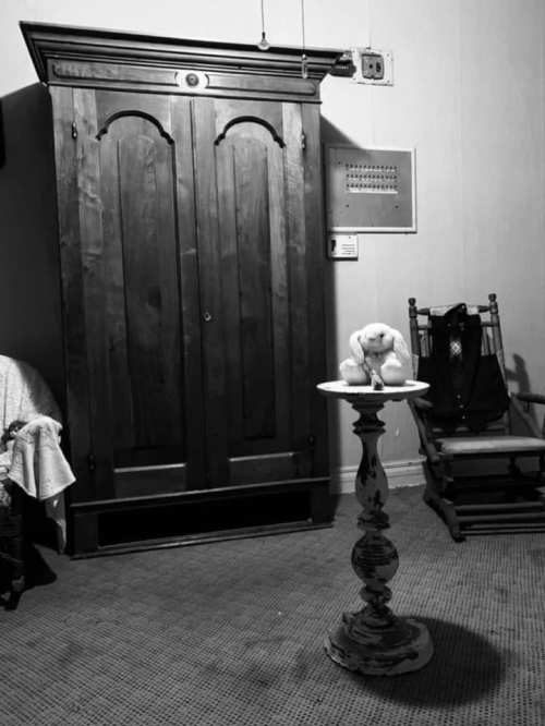 A vintage room featuring a wooden wardrobe, a small table with a stuffed animal, and a rocking chair.