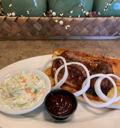 A plate with a sausage on a bun, topped with onions, alongside a side of coleslaw and barbecue sauce.