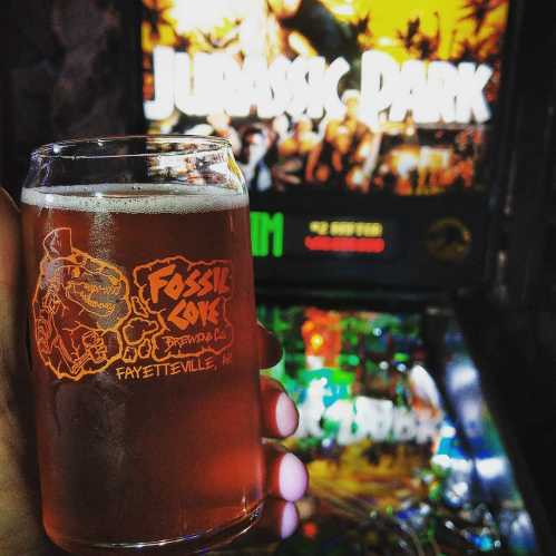 A hand holds a glass of amber beer in front of a Jurassic Park pinball machine.