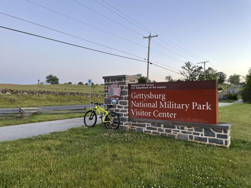 A bicycle leans against the sign for Gettysburg National Military Park Visitor Center, with grassy fields in the background.