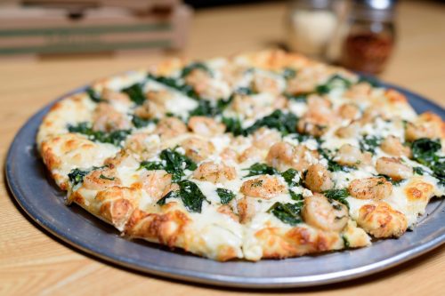 A close-up of a pizza topped with shrimp, spinach, and cheese on a metal tray, with pizza boxes in the background.