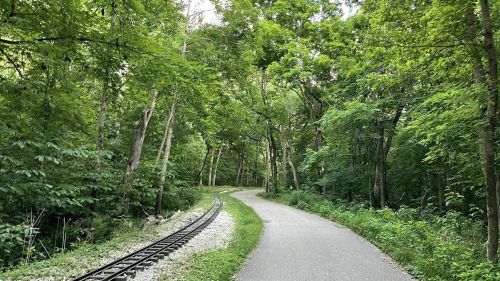A winding path through lush green trees, with a small railway track running alongside.