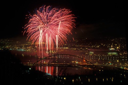 Colorful fireworks illuminate the night sky over a cityscape and bridge, reflecting in the water below.
