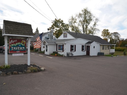 A white tavern building with a sign reading "Ron's Crooked Hill Tavern" and an American flag outside.
