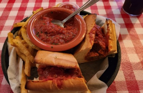 A plate with two meatball sandwiches, a bowl of marinara sauce, and a side of fries on a red and white checkered tablecloth.