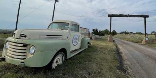 An old green truck parked beside a dirt road, with a wooden sign overhead and grassy fields in the background.