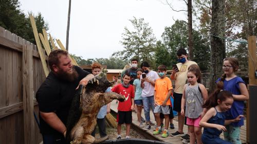 A group of children and adults watch as a man holds a large animal, surrounded by trees and wooden structures.