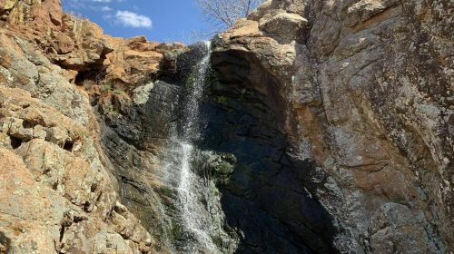 A small waterfall cascading down rocky cliffs, surrounded by textured stone and sparse vegetation under a blue sky.