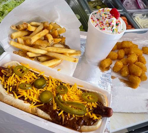 A loaded hot dog with cheese and jalapeños, served with fries, tater tots, and a milkshake topped with sprinkles and a cherry.