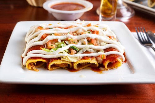 A plate of enchiladas topped with red sauce, lettuce, tomatoes, and drizzled with sour cream, served with a side of sauce.
