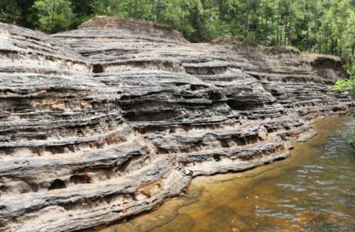 Layered rock formations beside a clear stream, surrounded by lush green trees in a natural landscape.