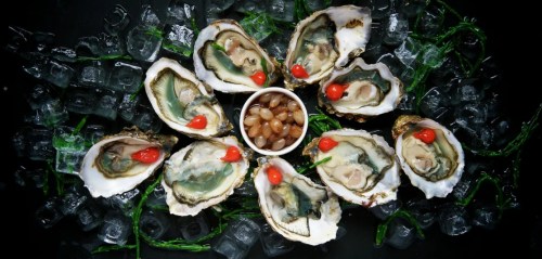 A platter of oysters on ice, garnished with red peppers and a small bowl of condiments in the center.