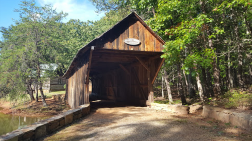 A rustic wooden covered bridge surrounded by trees, leading to a gravel path by a serene pond.