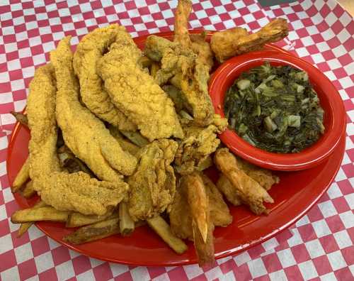 A red plate with fried fish fillets, shrimp, French fries, and a side of collard greens on a checkered tablecloth.