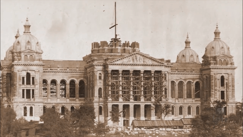 Historic building under construction, featuring scaffolding and a partially completed facade, with trees in the foreground.