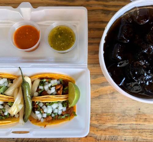 Two tacos with onions, cilantro, and jalapeños, served with two sauces and a cup of iced drink on a wooden table.