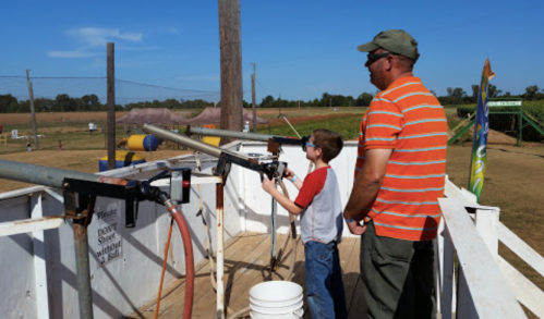 A man and a boy stand at a shooting range, aiming at targets with large guns on a sunny day.