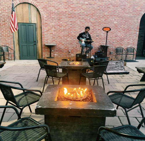 A musician plays guitar on a stage surrounded by fire pits and chairs in a cozy outdoor setting.
