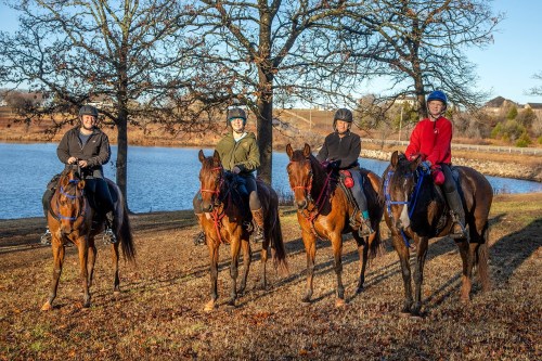 Four riders on horseback pose by a lake, surrounded by trees, with a clear blue sky in the background.