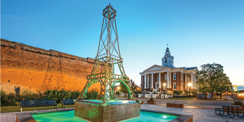 A green replica of the Eiffel Tower stands in a park with a fountain, beside a historic building at sunset.
