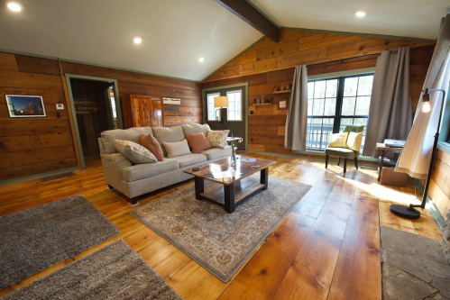 Cozy living room with wooden walls, a sofa, coffee table, and large windows letting in natural light.