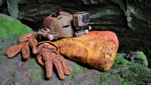 A weathered helmet and gloves resting on a mossy rock, surrounded by a cave-like environment.