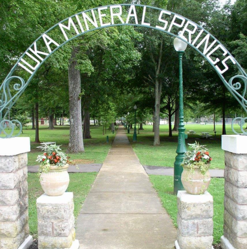 Archway reading "Iuka Mineral Springs" over a paved path lined with greenery and decorative planters.