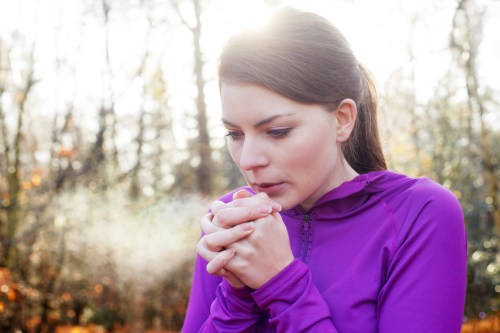 A woman in a purple jacket warms her hands, exhaling visible breath in a forest setting.