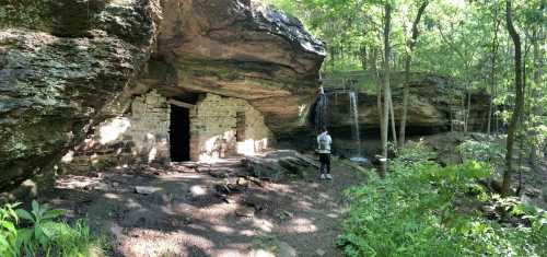 A person stands near a stone structure in a forest, with a small waterfall and lush greenery surrounding the area.