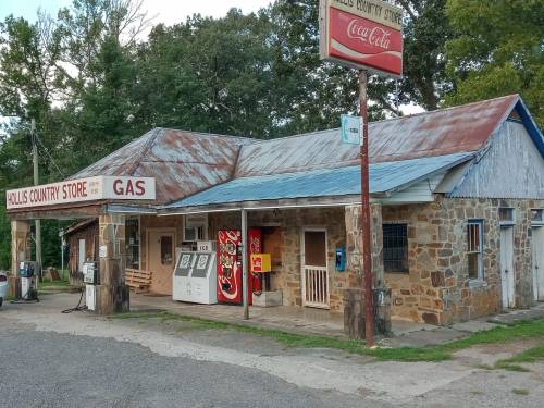 A rustic country store with a gas station, featuring a stone exterior and a Coca-Cola sign, surrounded by trees.