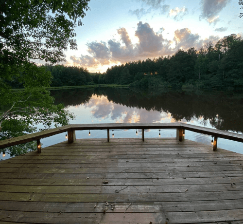 A wooden dock extends over a calm lake, surrounded by trees, with a colorful sunset reflecting on the water.