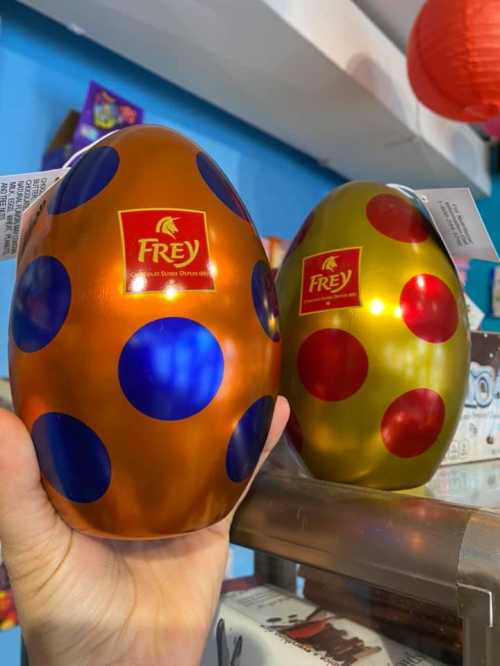 A hand holds two decorative chocolate eggs, one orange with blue dots and one gold with red dots, on a shelf.